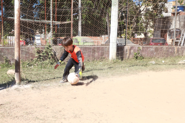Junge in Fußballschuhen auf einem Schotterfeld mit Gras, Pflanzen, Pfählen, einem Zaun, einer Wand, Bäumen, Fahrzeugen, Gebäuden und einem bewölkten Himmel im Hintergrund.