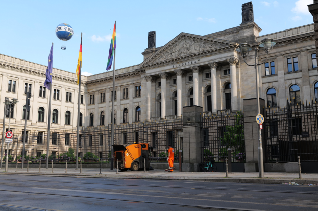 Großes modernes Gebäude mit vielen Fenstern, als Bundestag in Berlin identifiziert, umgeben von Straßeninfrastruktur, Fußgängern, Fahrzeugen, Flaggen und Bäumen unter einem bewölkten Himmel.