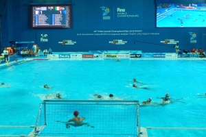 Eine Gruppe von Menschen, die Wasserball in einem großen Pool mit einem Netz in der Mitte spielen, Zuschauer sitzen auf Stühlen auf der linken Seite und eine Wand im Hintergrund zeigt Text und Logos für die FINA-Weltmeisterschaften 2016 in Kazan.