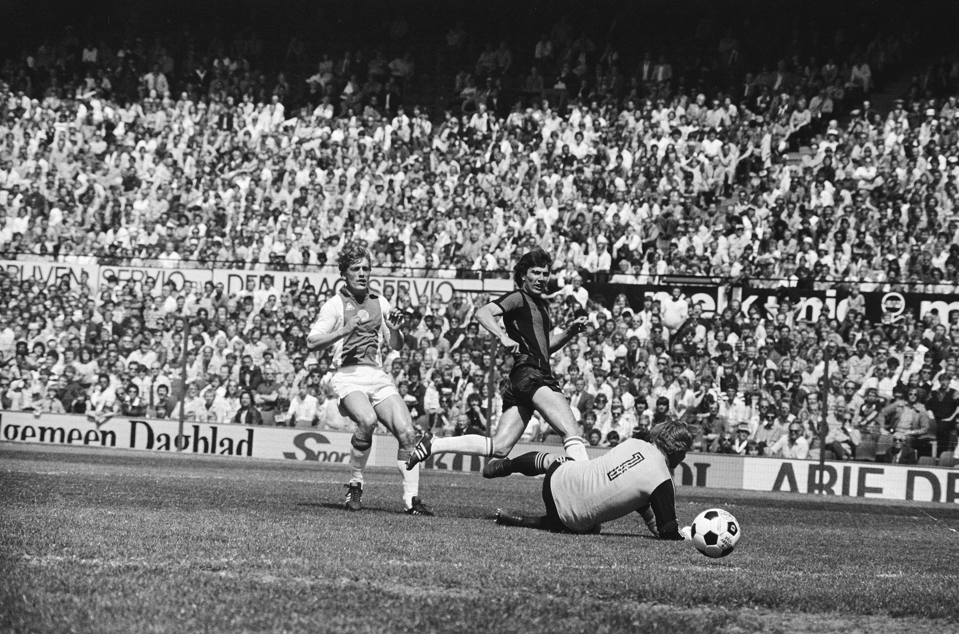 Schwarzes Foto von Männern, die Fussball auf einem Feld spielen, mit Zuschauern im Hintergrund und einer Fahne mit der Aufschrift 'Bundesliga 1978-79 Ajax 1-1 Hannover 96.'