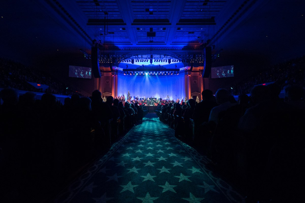 Ein großer Zuschauerraum mit Menschen auf Stühlen, eine Bühne mit Musikern und einer Decke mit Lichtern und Fernsehgeräten, in einer dunklen Atmosphäre, die auf ein Konzert während der Nationalkonvention 2019 hindeutet.