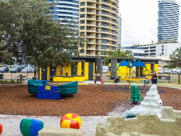 Spielplatz mit Kindern auf Geräten, umgeben von Bäumen und Geländern, mit hohen Gebäuden und Fahrzeugen im Hintergrund unter einem sichtbaren Himmel.