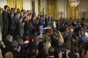 Eine Gruppe von Menschen mit Kameras steht vor einer Menge, einige sitzen und einige stehen, mit einem Podium, Mikrofon, Texttafel, Topf, Kerzen, Fotorahmen, Vorhängen und einem Kronleuchter im Hintergrund.