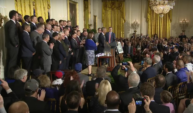 Eine Gruppe von Menschen mit Kameras steht vor einer Menge, einige sitzen und einige stehen, mit einem Podium, Mikrofon, Texttafel, Topf, Kerzen, Fotorahmen, Vorhängen und einem Kronleuchter im Hintergrund.