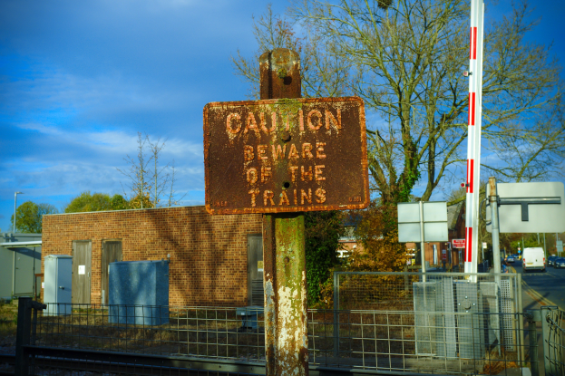 Warnschild an einem Bahnübergang-Zaun in der Nähe von Bäumen, Pfählen, einem Gebäude, Containern, Fahrzeugen und einem bewölkten Himmel.