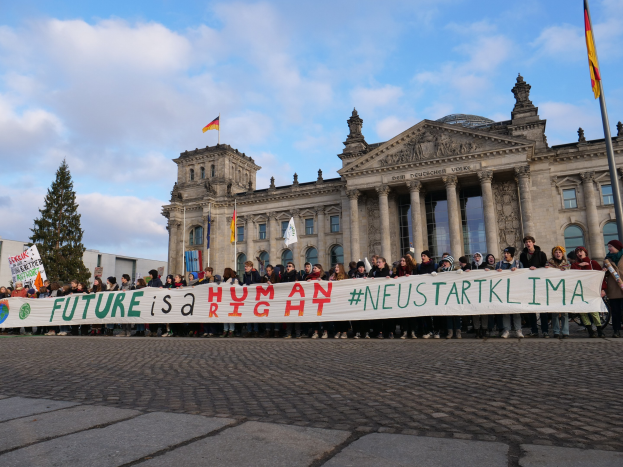 Eine Gruppe von Menschen hält ein "Zukunft ist ein Menschenrecht"-Plakat vor dem Reichstaggebäude in Berlin, mit Bäumen und Fahnenmasten im Hintergrund.
