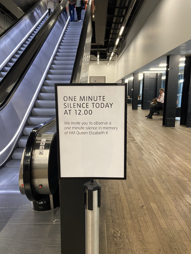 Eine Rolltreppe in einem Flughafen mit einem Schild, auf dem "Eine Minute Stille heute" steht, sowie ein paar Menschen darauf und an der Decke angebrachte Lichter im Hintergrund.