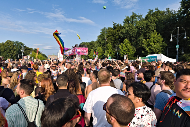 Große Menschenmenge vor einem Gebäude, einige halten Fahnen und Transparente, mit einem Zelt, Laternenmasten, Bäumen und einem Luftballon im Hintergrund, feiert die LGBT-Gemeinde.