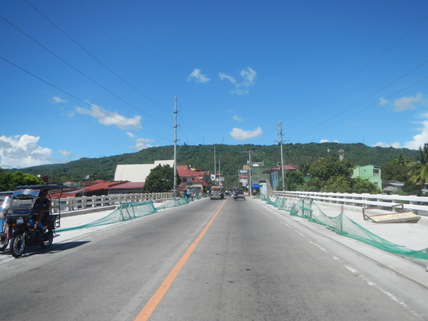 Autobahn mit fahrenden Fahrzeugen, einem geparkten Motorrad, Geländern, Strommasten mit Drähten, Bäumen, Gebäuden, Hügeln und einem bewölkten Himmel im Hintergrund.
