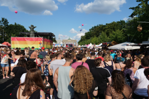 Eine große Menge Menschen geht eine Straße mit Zelten, Bäumen, Pfählen, Lichtern und einer Statue entlang, im Hintergrund sind Gebäude und ein Himmel mit Wolken und Ballons zu sehen während des Christopher Street Day Festivals in Berlin.