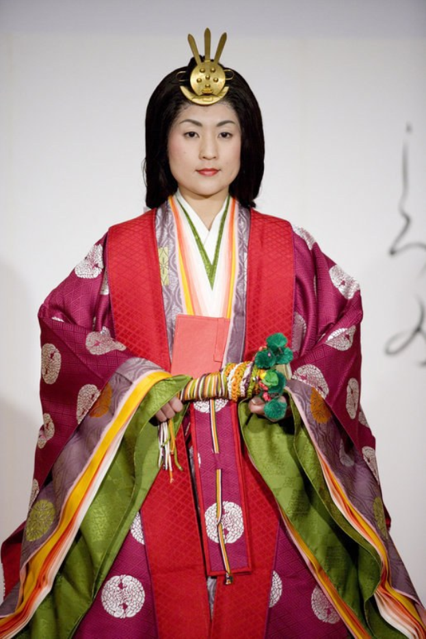 Eine Frau in einem traditionellen japanischen Kimono steht selbstbewusst auf einem Laufsteg und trägt ein leuchtend rotes und grünes Kleid mit kunstvollen Mustern, ihr Haar ist ordentlich zu einem Knoten gebunden.
