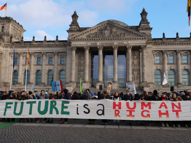 Eine Gruppe von Menschen hält ein Transparent mit der Aufschrift "Zukunft ist ein Menschenrecht" vor dem Reichstaggebäude in Berlin, Deutschland, mit sichtbaren Fenstern, Säulen und Bögen sowie umgeben von Fahnen mit Stangen unter einem bewölkten Himmel.