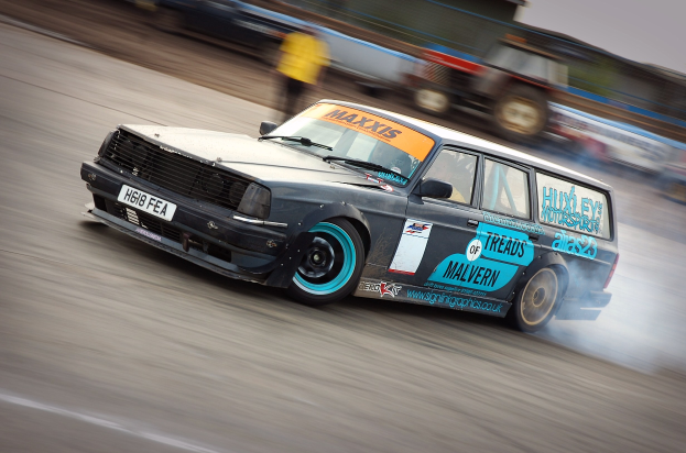 Ein Auto fährt eine Rennstrecke hinunter, wobei Rauch ausstößt, mit anderen Fahrzeugen und Menschen im Hintergrund.