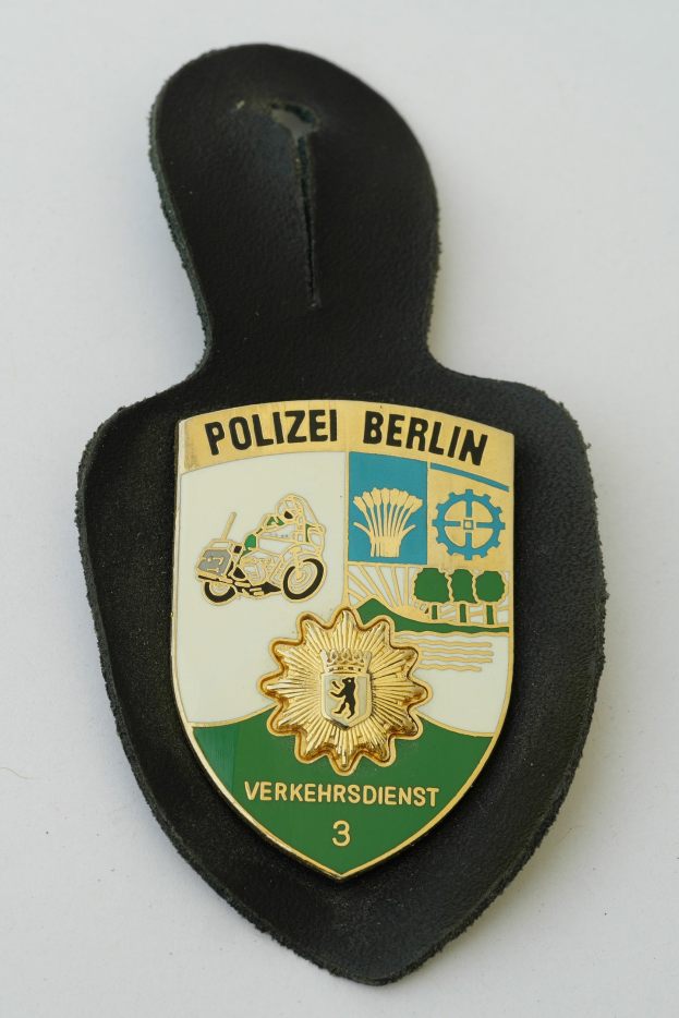 Ein schildförmiges Polizeisiegel mit 'Polizei Berlin' in schwarzer, fetter Schrift auf einem blauen Hintergrund mit einem weißen Stern und Rahmen, das auf einer Oberfläche ruht.