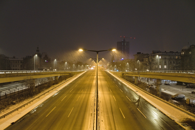 Nachtszene auf der Autobahn mit fahrenden Autos, Straßenlaternen, Geländern, Bäumen, Gebäuden mit beleuchteten Fenstern und sichtbarem Himmel.