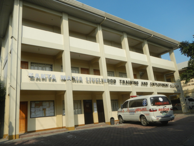 Außenansicht des Santa Maria Lebensmittel-Trainings- und Beschäftigungszentrums mit Fenstern, Türen und Beschriftung, umgeben von Bäumen und Fahrzeugen auf der Straße unter einem klaren blauen Himmel.