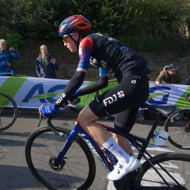 Professionelle Radrennfahrerin Katie Archibald trägt Helm, Brille, Handschuhe und Schuhe und fährt ein Fahrrad an einer Zuschauermenge vorbei, mit einem Banner, einer Wand, Bäumen und einem Gebäude im Hintergrund.