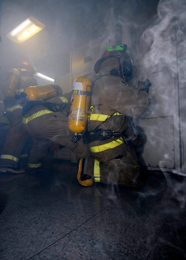 Feuerwehrleute in Schutzausrüstung mit Rauch, der aus einer Türschwelle quillt, beleuchtet von einem Licht an einer Wand.