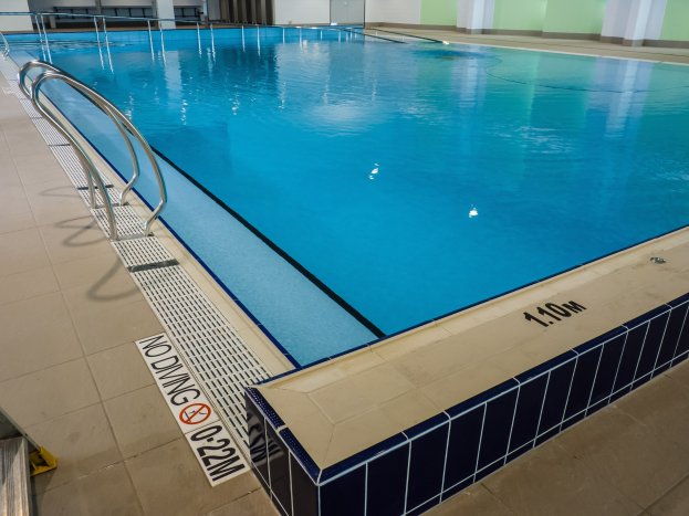 Großes Hallenbad in einem geräumigen Gebäude mit gefliestem Boden, Metallträgern, einer Tafel, einer Tür, einer Wand und Deckenlampen.
