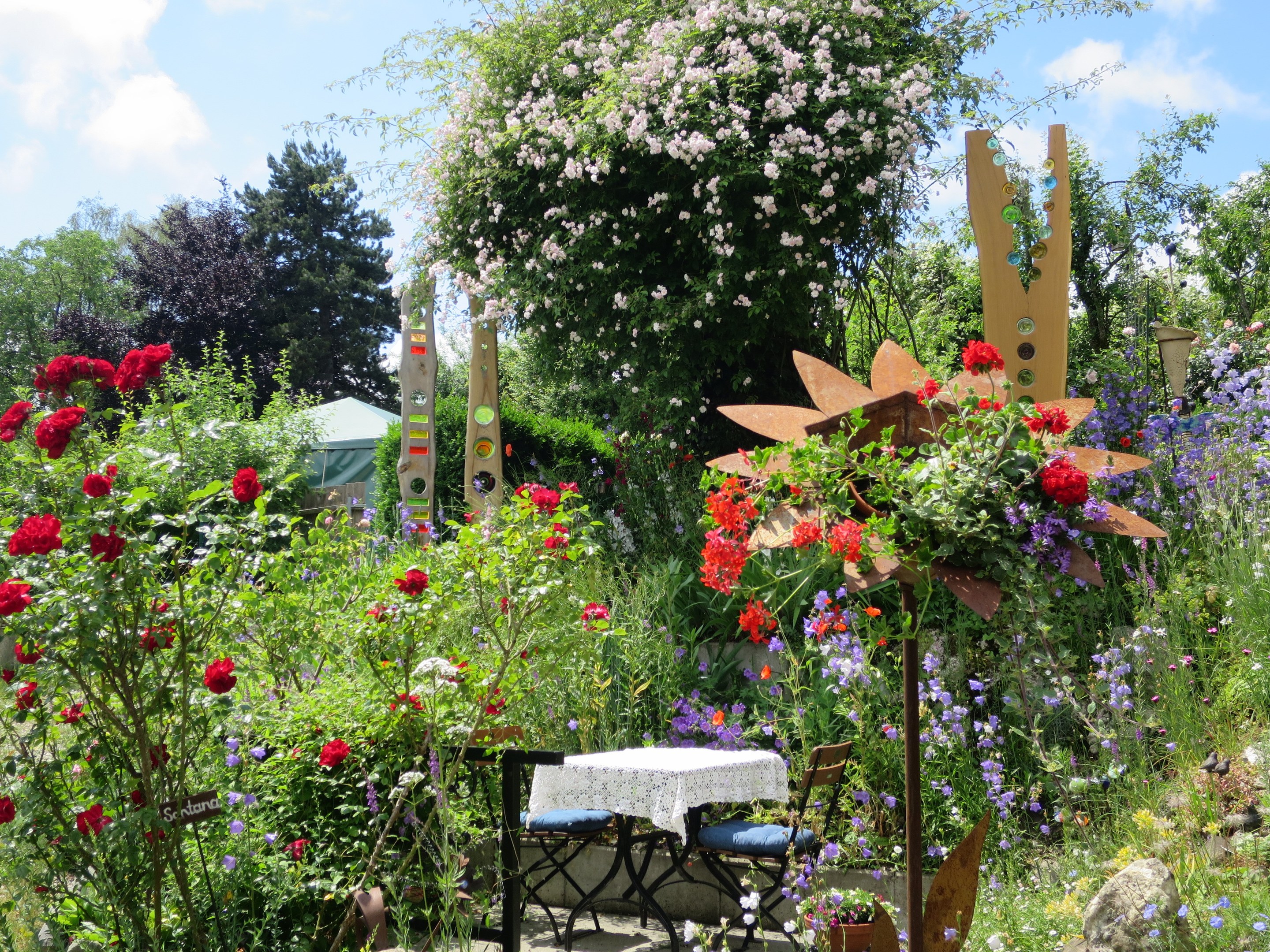 Ein Garten mit bunten Blumen und Pflanzen, der einen Tisch und Stühle mit einer Decke zeigt, vor Bäumen und einem bewölkten Himmel auf der Chelsea Flower Show.