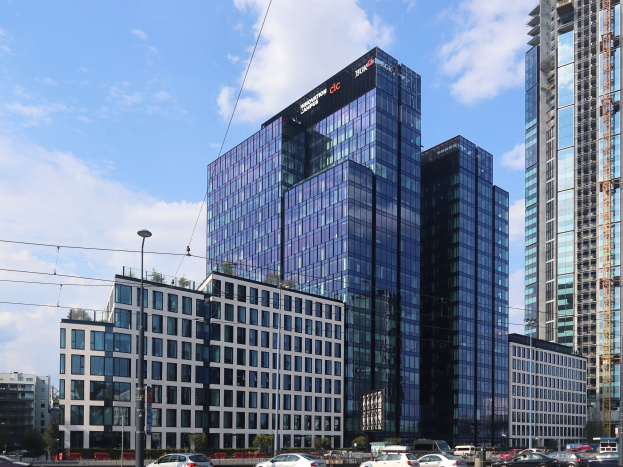 Großes modernes Gebäude mit zahlreichen Fenstern, identifiziert als die Deutsche Bank Hauptverwaltung in Frankfurt, Deutschland, umgeben von städtischer Infrastruktur einschließlich Straßenlaternen, Fahrzeugen, Bäumen und einem bewölkten Himmel.