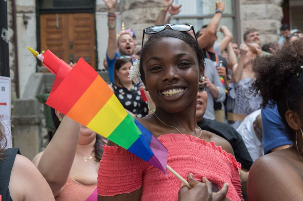 Eine Gruppe von Menschen vor einem Gebäude mit einer Frau in der Mitte, die eine Regenbogenflagge hält, wahrscheinlich an einem Christopher Street Day teilnehmend.