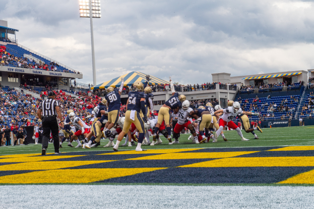 Ein Fussballspiel im Gange mit Spielern in Sportbekleidung und Helmen im Vordergrund, Zuschauern, Geländern, Beschriftungen, Laternen und einem bewölkten Himmel im Hintergrund und Text unten.
