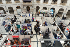 Große Besuchergruppe auf einer internationalen Fachmesse mit Stönden, Bannern, Tischen, Stühlen und Luftballons in einem Raum mit architektonischen Bogengängen und Pfeilern.