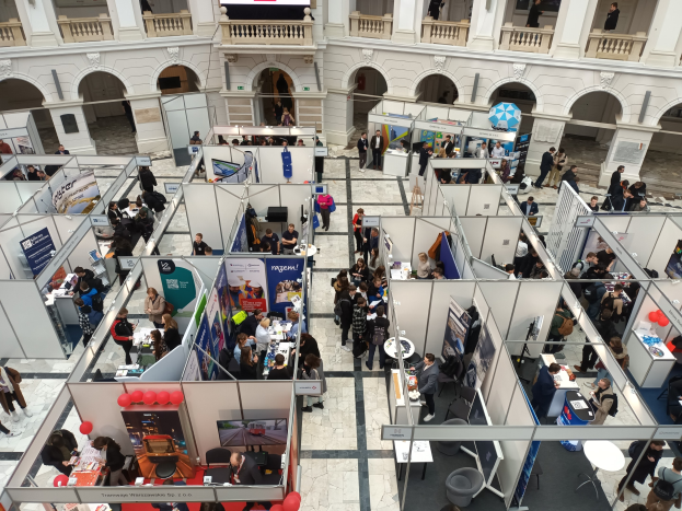 Große Besuchergruppe auf einer internationalen Fachmesse mit Stönden, Bannern, Tischen, Stühlen und Luftballons in einem Raum mit architektonischen Bogengängen und Pfeilern.