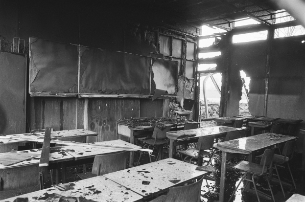 Schwarz-weiß-Foto eines verlassenen Klassenzimmers mit verkohlten Wänden, zerbrochenen Fenstern und verstreuten Tischen und Stühlen.