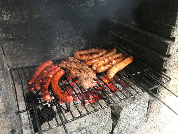 Grill mit Würstchen und Fleisch darauf, umgeben von Kohle und Feuer, auf Gras im Freien