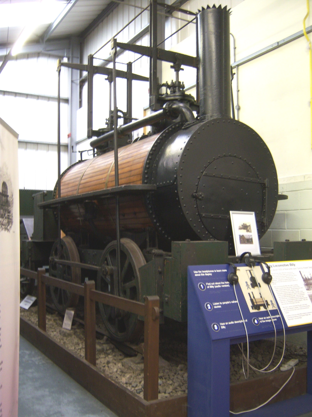 Alte Dampflokomotive in einem Museum ausgestellt, umgeben von Informationsschildern, einem Zaun und Deckenleuchten.