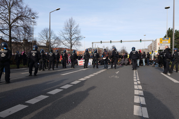 Eine Gruppe von Polizisten in schwarzen Uniformen und Helmen, die an der Seite einer Straße mit Laternenmasten, Ampeln, Bäumen, Gebäuden und einem klaren blauen Himmel im Hintergrund stehen.