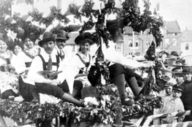 Schwarzes und weißes Bild eines Paradewagens, der Menschen in traditioneller bayrischer Tracht trägt, einige spielen Instrumente, mit Blattdekorationen vor einem Hintergrund aus Gebäuden und einem klaren Himmel.