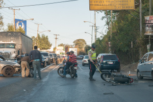 Eine Gruppe von Menschen umringt ein verunglücktes Motorrad am Straßenrand mit mehreren Fahrzeugen, darunter ein Lastwagen, und einem Hintergrund aus Bäumen, Polen, Lampen und Schildern unter dem Himmel.
