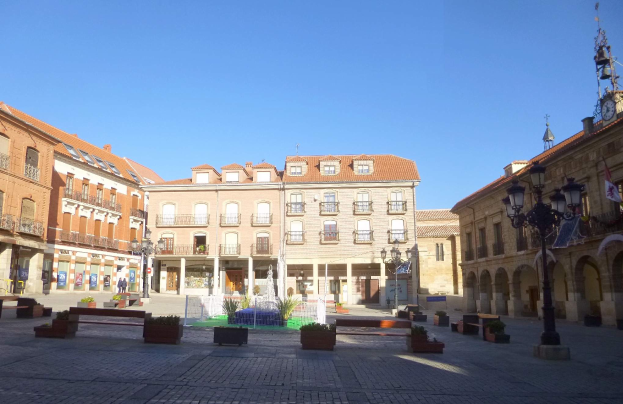 Ein Plaza Mayor in einem Stadtplatz mit einem zentralen Brunnen, umgeben von Bänken, Topfpflanzen, Straßenlaternen, einem Uhrenturm und Gebäuden mit Fenstern unter einem klaren blauen Himmel.