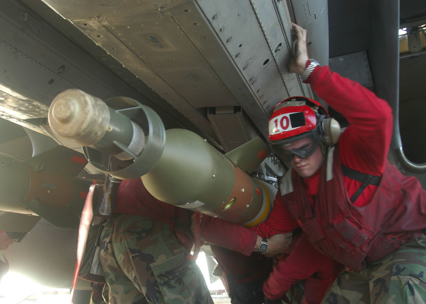 Eine Gruppe von Männern in roten Jacken und Tarnuniformen arbeitet an einem Flugzeug, wobei einer einen Helm, eine Schutzbrille und eine Uhr trägt und im Hintergrund eine Rakete zu sehen ist.