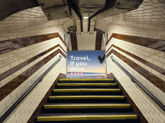Eine Treppe in einer U-Bahn-Station mit einem Schild oben, das 'Reise, wenn du wagst' lautet, flankiert von Geländern und sichtbaren Stationwänden.
