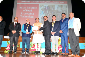 Eine Gruppe von Männern auf einer Bühne mit einem Zertifikat und einer Projektion im Hintergrund, die 'Indian Institute of Food Processing Technology Awards 2019' und Blumenarrangements zeigt.