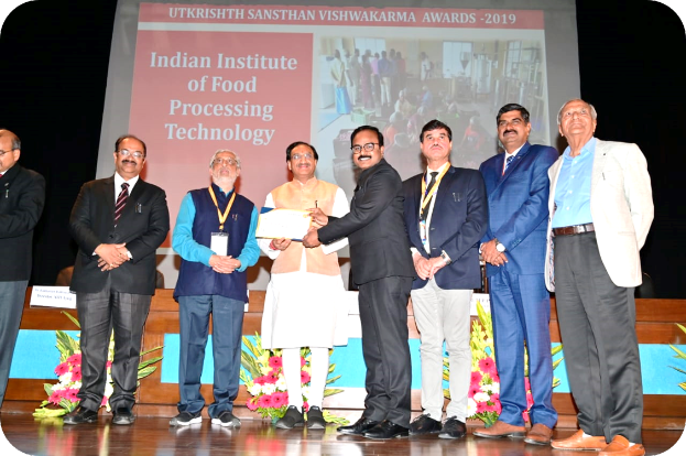 Eine Gruppe von Männern auf einer Bühne mit einem Zertifikat und einer Projektion im Hintergrund, die 'Indian Institute of Food Processing Technology Awards 2019' und Blumenarrangements zeigt.