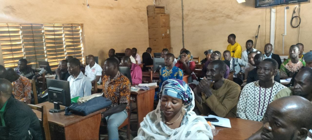 Eine Gruppe von Menschen sitzt an Tischen in einem Klassenzimmer mit Monitoren, Taschen, Büchern und Papieren und nimmt an einem Techniktraining teil, mit einem Fenster auf der linken Seite und Kartons und einem Fernseher an der Wand im Hintergrund.
