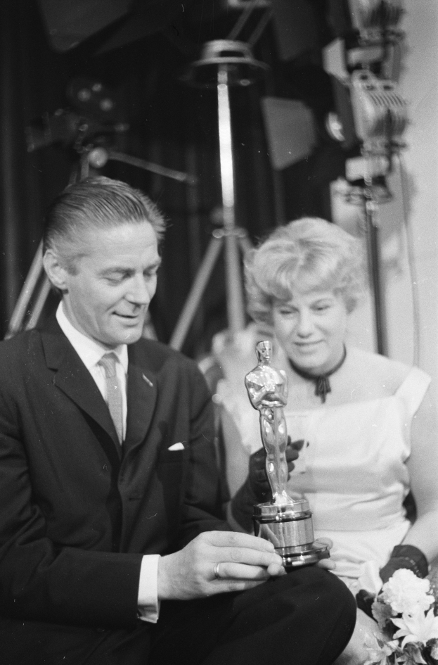 Ein Mann in einem schwarzen Blazer, weißem Hemd und Krawatte hält eine Oscar-Statue, während er neben einer Frau in Abendgarderobe sitzt, die einen Blumenstrauß hält, mit Hintergrundbeleuchtung, die die schwarz-weiße Szene erhellt.