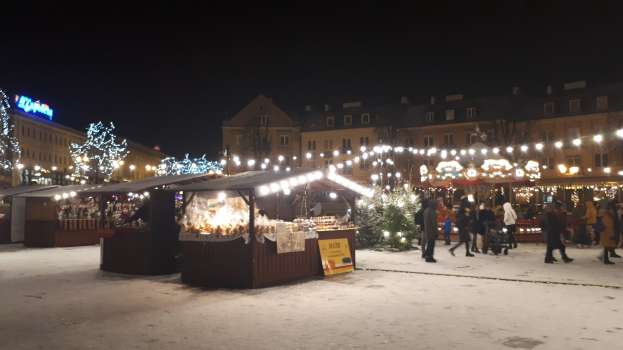 Ein geschäftiges Weihnachtsmarkt in einer verschneiten Nacht mit Menschen, Buden mit Lichtern, Pflanzen, Bäumen, Gebäuden und Schildern unter einem bewölkten Himmel.