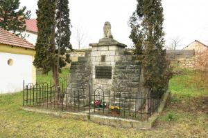 Ein steinerner Gedenkstein auf einer Rasenfläche, umgeben von einem Metallzaun und geschmückt mit Blumensträußen, mit Bäumen, Häusern und einer Wand im Hintergrund unter einem klaren blauen Himmel, als Holocaust-Gedenkstätte in Sibiu, Rumänien.