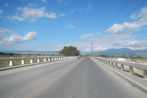 Eine Autobahn mit Motorradfahrern, umgeben von Geländern, Strommästen mit Käblein, Bäumen, Hügeln und einem bewölkten Himmel.