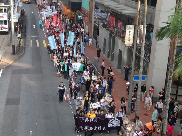 Eine große Gruppe von Menschen marschiert durch eine Straße in Hong Kong, mit Protestschildern in den Händen, mit Bäumen und Glasgebäuden auf der rechten Seite und Fahrzeugen auf der linken Seite.