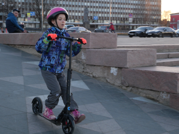 Ein junger Junge in Helm und Handschuhen fährt mit einem Roller eine Treppe hinunter, mit Fahrzeugen, Menschen, Bäumen, Polen, Brettern, Gebäuden und einem klaren blauen Himmel im Hintergrund.