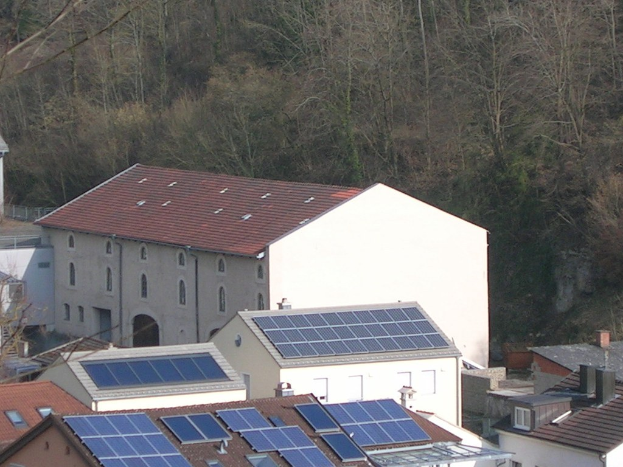 Haus mit Solarpaneelen auf dem Dach, umgeben von anderen Gebäuden und Bäumen im Hintergrund.