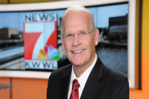 Ein Mann im Anzug und Krawatte, mit Brille, steht lächelnd vor einem Fernseher, mit einem unscharfen Hintergrund.