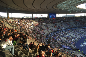 Eine große Menschenmenge sitzt im Allianz Stadion in München, Deutschland, bei einem Fussballspiel, mit einer Bühne auf der rechten Seite, Fahnen, Mästängen und einem Bildschirm im Hintergrund und dem Himmel oben sichtbar.